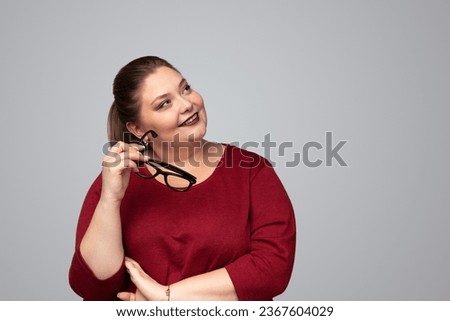 Positive young smart plus size woman with long dark hair and red lips in casual clothes, looking away dreamily with eyeglasses in hand against gray background Royalty-Free Stock Photo #2367604029