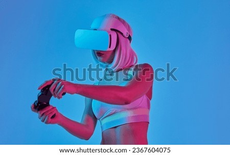 Futuristic young female in modern VR headset using controller to play video game while standing under neon light against blue background