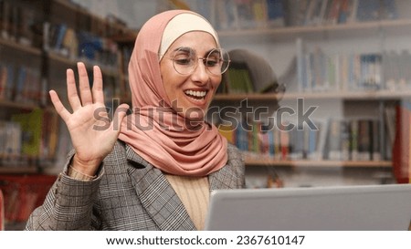 Portrait of a cute smiling Muslim girl in hijab and glasses sitting in the library against the background of bookshelves and looking at the computer screen, waving her hand in greeting. Online meeting