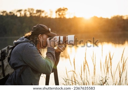 Wildlife photographer with camera photographing nature on lake at sunset. Man is taking picture outdoors