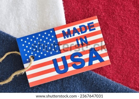 Made in USA label sitting on top of red, white and blue clothing shirts as a business economics concept. Flat lay macro image with patriotic background details. Royalty-Free Stock Photo #2367647031