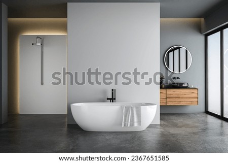 Modern bathroom interior with blue and white tones wall, concrete floor, wooden vanity with black sink and oval mirror, white bathtub, panoramic windows. Royalty-Free Stock Photo #2367651585