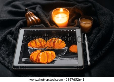 The still life picture with pumpkins drawn on the electronic tablet next to burning candle and ceramic pumpkin figurine on black table. The concept of inspiration, creativity, self-development, hobby,