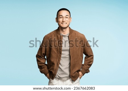 Portrait of attractive smiling Asian man wearing stylish brown jacket and eyeglasses, hands in pockets posing isolated on blue background. Advertisement concept Royalty-Free Stock Photo #2367662083