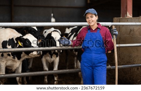Young female farmer in overalls posing while feeding cows at dairy farm Royalty-Free Stock Photo #2367672159
