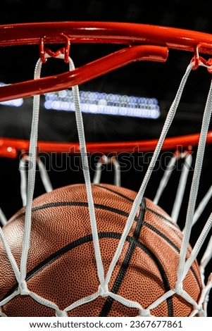 A close-up view looking up at the top of an orange basketball falling through the rim and a white nylon net with the arena lights and lens flare.