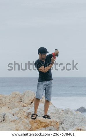 Indonesian male photographer wearing a hat and black t-shirt standing on coral rocks with a beach view in the background. Outdoor active vacation adventure travel concept.