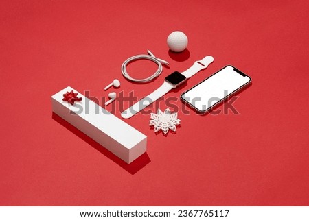 Christmas concept flat lay on red background with white objects. Blank screen smart phone mockup, smart watch, earbuds, gift box, charging cable and Christmas ornament.