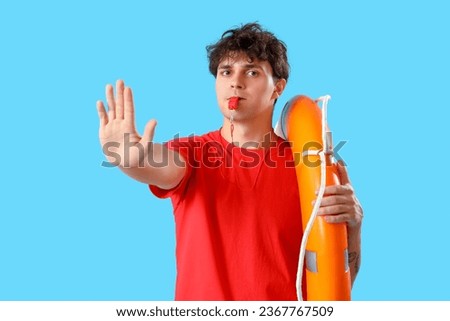 Male lifeguard with ring buoy and whistle showing stop gesture on blue background Royalty-Free Stock Photo #2367767509