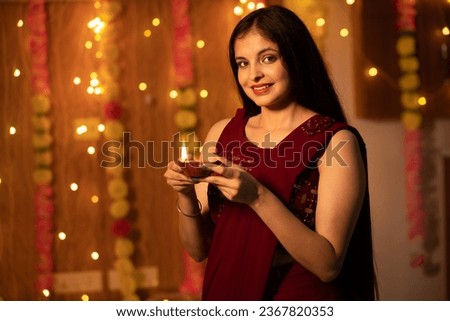Portrait of a beautiful young smiling Indian woman in traditional dress holding oil lamp light or diya with festive lights decoration in the background celebrating diwali.
