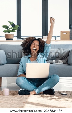 Shot of beautiful woman celebrating something while working with laptop sitting on the home Royalty-Free Stock Photo #2367834439