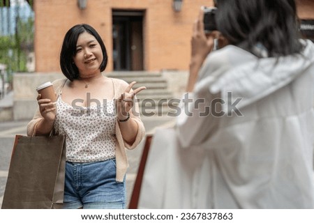 A happy Asian plus-size woman is being taken a photo by her friend while enjoying shopping day in the city together, carrying shopping bags, showing the peace hand sign and smiling at the camera.