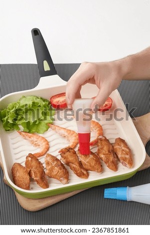     Close-up shot of a man applying oil on meat with silicone brush. Meat, shrimp and vegetables lying on the grill pan. A man's hand uses a silicone oil bottle with basting brush. Top view.           Royalty-Free Stock Photo #2367851861