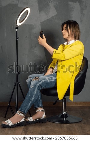 Brunette with short hair taking selfie leaning back on black leather chair near annular LED lamp. Young lady in yellow coat and blue jeans looking at phone screen sitting against gray wall. Vertical.
