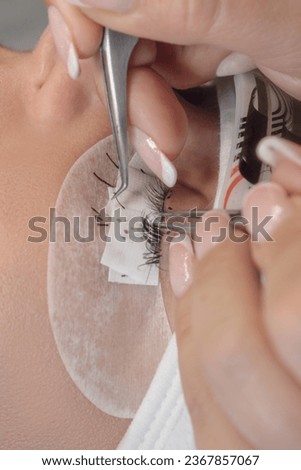 Lashmaker with tweezers in hands parting cilia for sticking black artificial eyelash on eyelid of client with closed eyes and patches on lower lid. Master attaching fake lashes in place. Vertical. Royalty-Free Stock Photo #2367857067