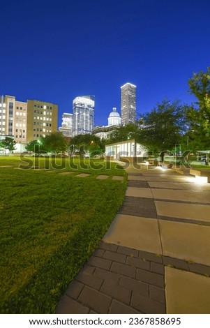 Illuminated pathway concrete sidewalk at park square with downtown Houston skyscraper, capitol tower in background during evening blue hour. Modern high rise cooperates office buildings metro complex