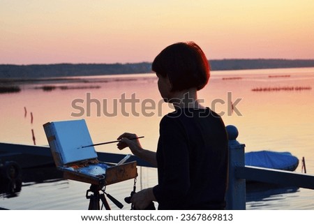 A girl Artist paints a sunset on Lake Pleshcheyevo on canvas at dusk