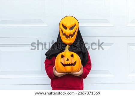 A child girl wearing a scary Halloween pumpkin mask, holding a pumpkin, over a white garage door. Concept of celebration, costume, carnival, terror, fear and autumn.