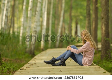 Sad young adult woman sitting alone on wooden trail at birch tree forest in autumn day. Thinking about life problem at nature park. Suffering from relationship break up. Side view. Royalty-Free Stock Photo #2367889703