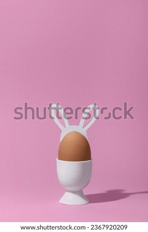 Brown egg with hare ears in a white eggcup on pink background. Minimal easter concept.