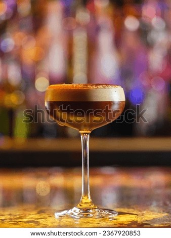 Glass full of brown alcoholic espresso cocktail with white froth served in bar with bottles against blurred background with bokeh