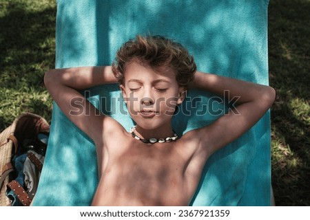 From above of shirtless boy with blond hair lying on blue blanket with hands clasped behind head while relaxing in garden with eyes closed