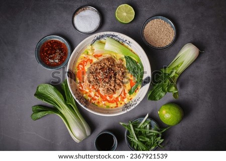 Stock photo of yummy noodles soup with minced meat isolated in grey background next to sesame seeds and other ingredients.