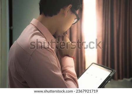 The image of a young man sitting and contemplating his work on an iPad in a room with little sunlight streaming through the curtained open window.
