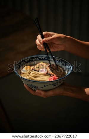 Crop anonymous female with chopsticks eating yummy ramen soup with noodles and meat in bowl against dark background