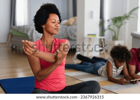 Young woman, mother exercising at home in living room, father playing with kid in background.