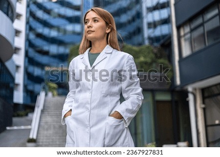 Female doctor walking outdoors in front of hospital. Hospital worker standing outside hospital while smiling and looking away. Royalty-Free Stock Photo #2367927831