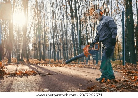 Professional maintenance worker in uniform cleaning city park from dry fall leaves with hand blower. Side view of caucasian aged man taking care of city area during autumn time. Work, season concept.