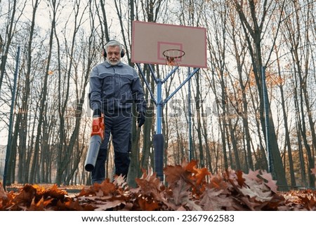 Aged janitor taking care of park area during autumn season. Low angle view of bearded man in overalls using electric blower to remove dry leaves at outdoors basketball court. Seasonal work concept.