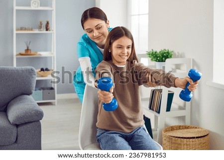 Child girl doing exercise using dumbbells with support from nurse.Young smiling caregiver physiotherapist helping teenage kid in lifting dumbbell for rehab. Healthcare medical concept.