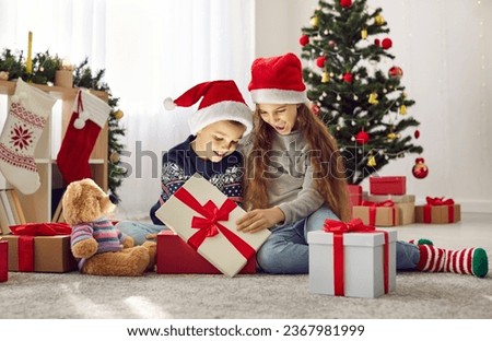 Christmas miracle. Cheerful little boy and girl in red Santa Claus hats happily open Christmas presents in morning. Kids look with interest at gift from which light shines and illuminates their faces.