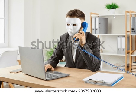 Anonymous man disguised in white face mask sitting at office desk with laptop, holding telephone receiver, making phone call, trying to scam people and steal their money. Phone fraud concept Royalty-Free Stock Photo #2367982003