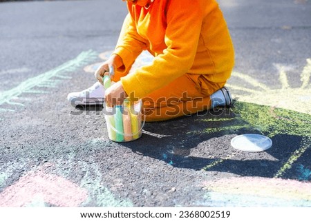 A little seven-year-old boy in an orange tracksuit and white sneakers draws with colored chalk on the asphalt
