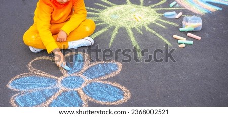 A little seven-year-old boy in an orange tracksuit and white sneakers draws a flower with colored chalk on the asphalt