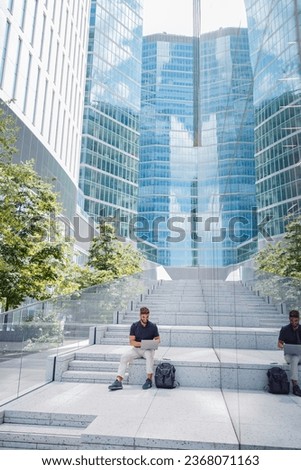Smiling bearded male freelancer working on laptop sitting on skyscrapers background