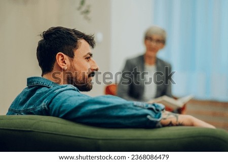 Profile of a young man with issues sitting on a sofa in the office with a psychotherapist and discussing trauma and problems. In a blurry background is the therapist giving support and giving advice. Royalty-Free Stock Photo #2368086479