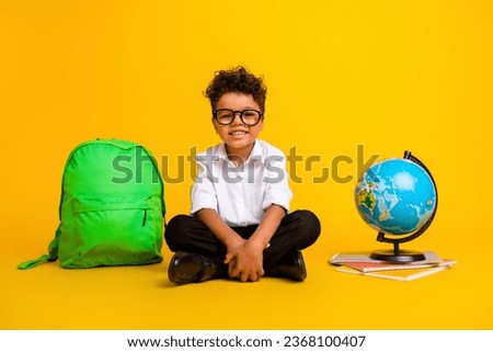 Full length photo of little intelligent schoolkid sitting floor rucksack world globe copybook isolated on yellow color background
