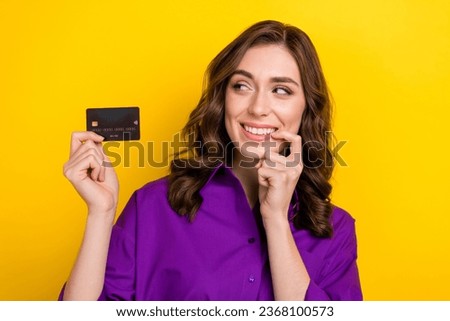 Photo of minded intelligent woman with wavy hair dressed purple shirt look at plastic card finger on teeth isolated on yellow background