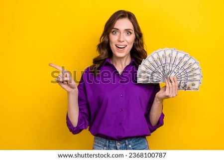 Photo of impressed excited woman with curly hairdo dressed purple shirt hold dollars indicating empty space isolated on yellow background