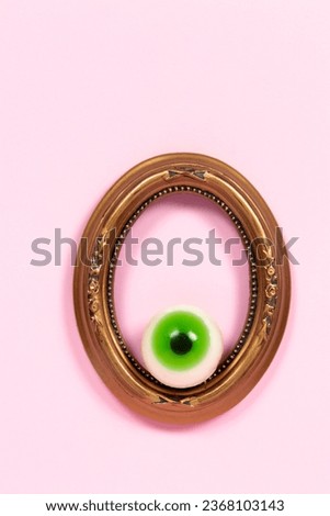 Green candy eye in an old painting frame on pastel pink background. Halloween concept.