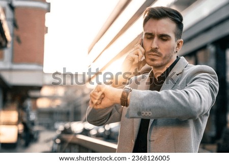 Handsome businessman talking on the cell phone while looking at his wristwatch in urban city street. Busy male person arranging a business meeting over phone.