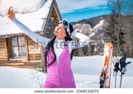 Young beautiful female skier taking a selfie in front of mountain cottage. Attractive young woman with black long hair standing outdoors on a sunny winter day and posing for self portrait.
