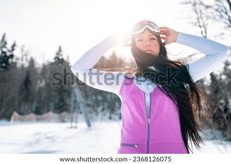 Portrait of a beautiful young female snowboarder looking at camera. Waist up photo of attractive girl with long hair enjoying winter holiday outdoors.