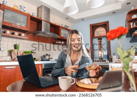 Cheerful blonde woman sitting at table and eating breakfast in kitchen. Smiling girl is holding knife and spreading cream cheese on her bread.