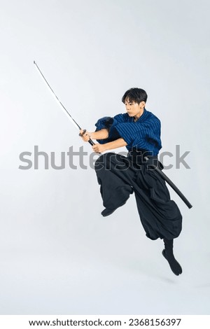 Japanese young samurai wielding katana sword while jumping in studio with white background. Royalty-Free Stock Photo #2368156397