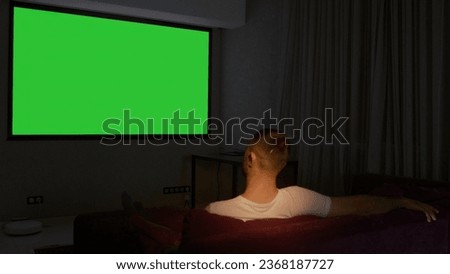A man sitting on the couch. Media. An adult male resting in a room looking at a green screen.